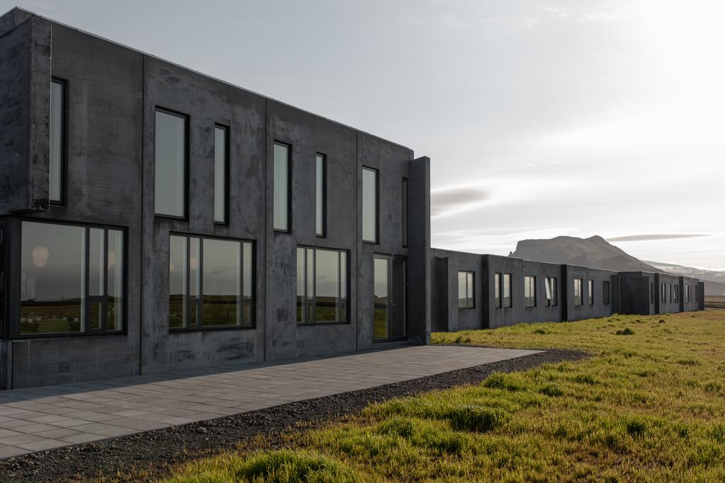 Photo of the exterior of Umi Hotel located in Vik Iceland. The building is dark gray and very rectangular. Mountains are seen in the distance.