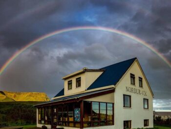 Photo of Vik Hostel in Vik Iceland. A pale yellow farmhouse is seen with a dark blue roof. Picnic tables are located outside of the building. A rainbow is right above with stormy clouds.