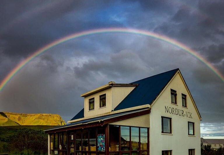 Photo of Vik Hostel in Vik Iceland. A pale yellow farmhouse is seen with a dark blue roof. Picnic tables are located outside of the building. A rainbow is right above with stormy clouds.