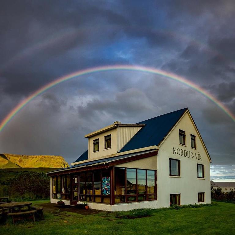 Photo of Vik Hostel in Vik Iceland. A pale yellow farmhouse is seen with a dark blue roof. Picnic tables are located outside of the building. A rainbow is right above with stormy clouds. 