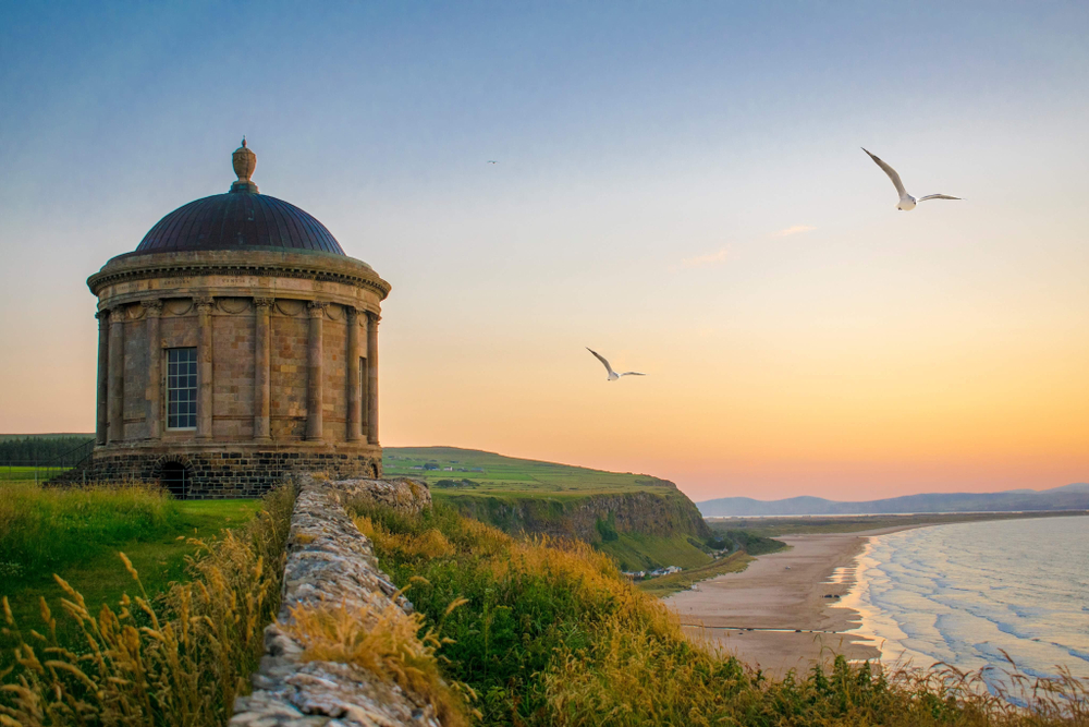 Mussenden Temple is one of the most photographed places in Ireland so should definitely be on your 'things to do in Northern Ireland' list. The photo shows the circular temple on the edge of a cliff. There is a beach in the background and a gorgeous sunset. There are birds flying in the sky.