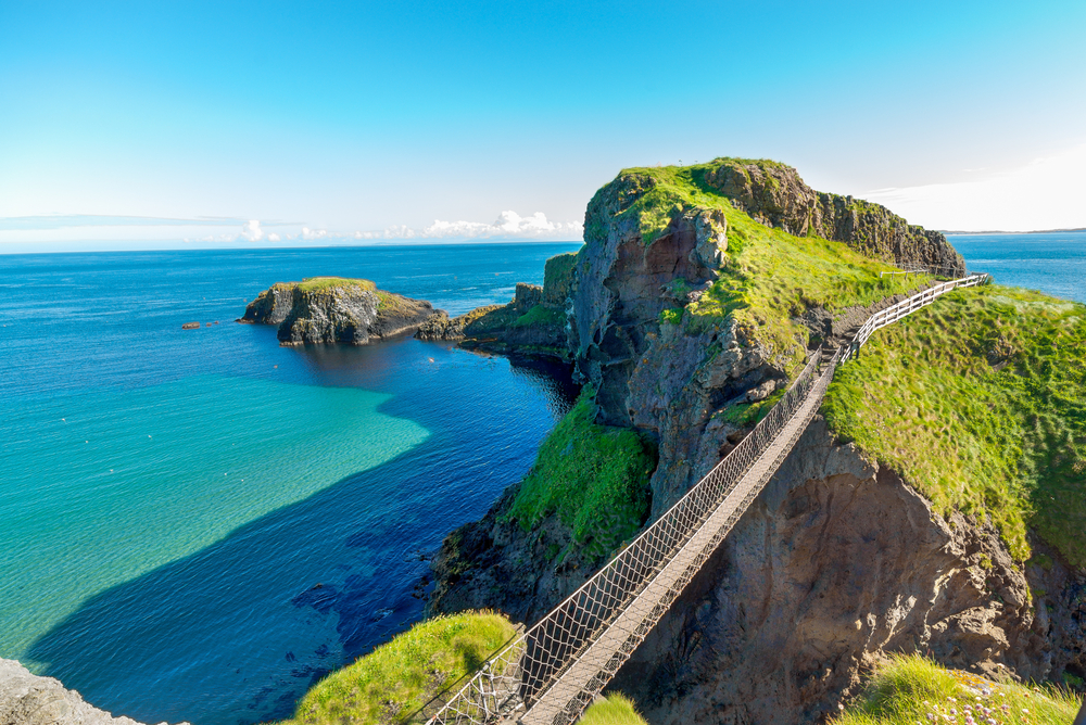Birds-eye-view of the Carrick-a-Rede rope bridge between rugged cliffs on a sunny day.