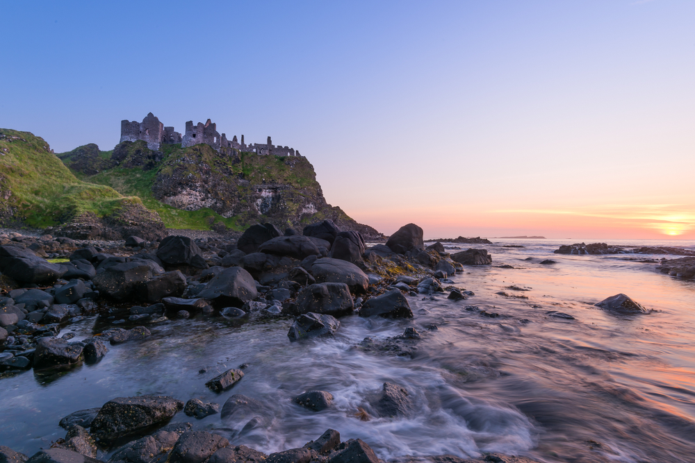 sunset view of Dunluce Castle in Northern Ireland. The castle is in the background and a rocky beach is in the foreground