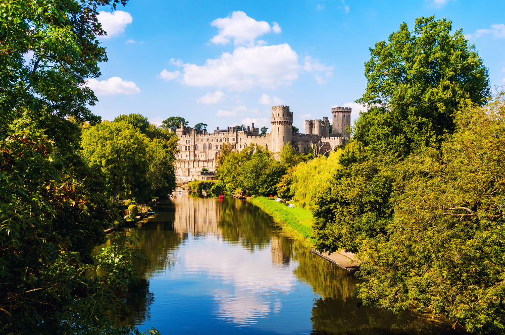 a photo of a lake with the beautiful Warwick Castle in the background, surrounded by trees.