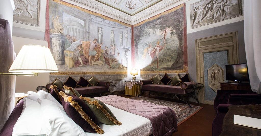 Photo of guest room at Hotel Burchianti in Florence Italy. Room is ornate a large bed with white linen sheets and plum and green velvet pillows. Plum velvet cushioned benches line the room. Large frescoes are on the walls and ceiling.