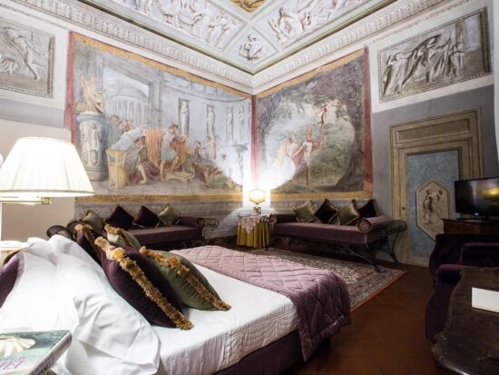 Photo of guest room at Hotel Burchianti in Florence Italy. Room is ornate a large bed with white linen sheets and plum and green velvet pillows. Plum velvet cushioned benches line the room. Large frescoes are on the walls and ceiling.