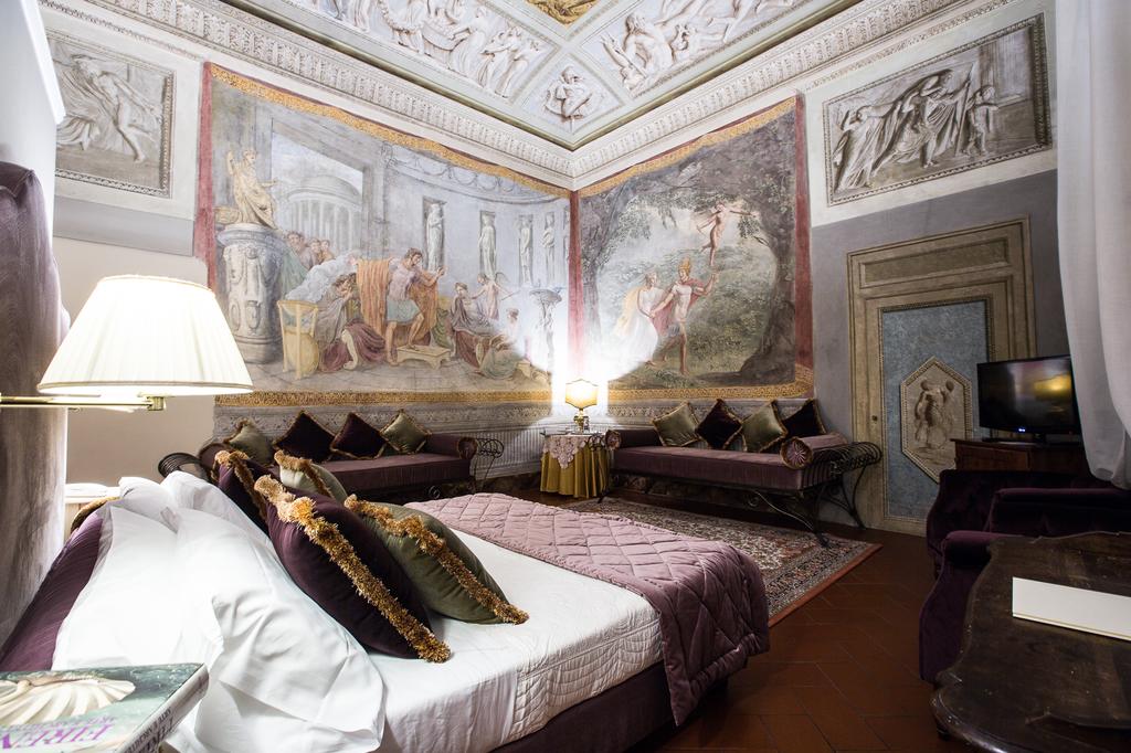 Photo of guest room at Hotel Burchianti one of the best places to stay in Florence. Room is ornate a large bed with white linen sheets and plum and green velvet pillows. Plum velvet cushioned benches line the room. Large frescoes are on the walls and ceiling. 