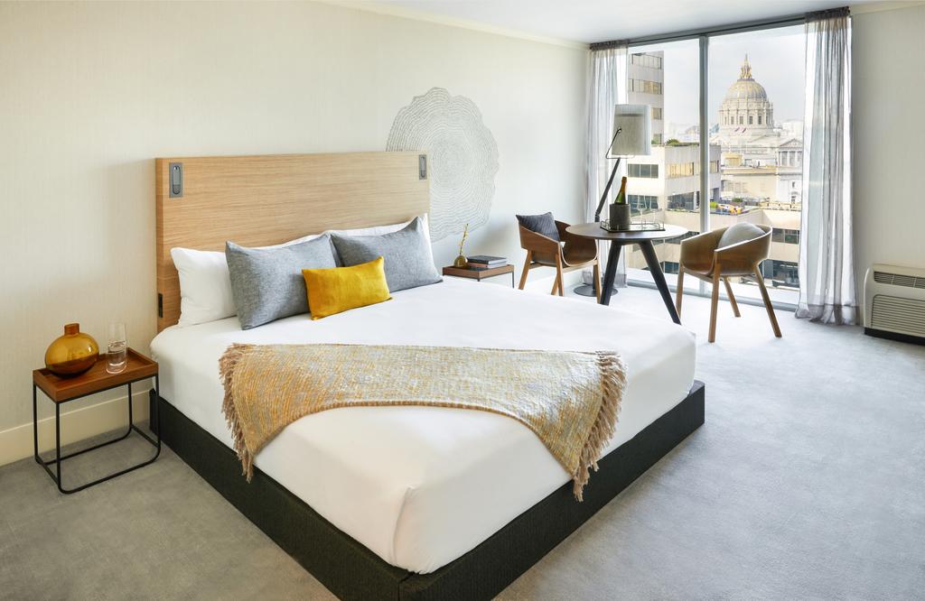 Photo of guest room at BEI Hotel located in Downtown San Francisco which is one of the best places to stay in San Francisco. A guest bed is seen with white linens and a tan throw blanket. Multi-colored throw pillows are arranged on the bed. A window balcony is seen with a city view. 