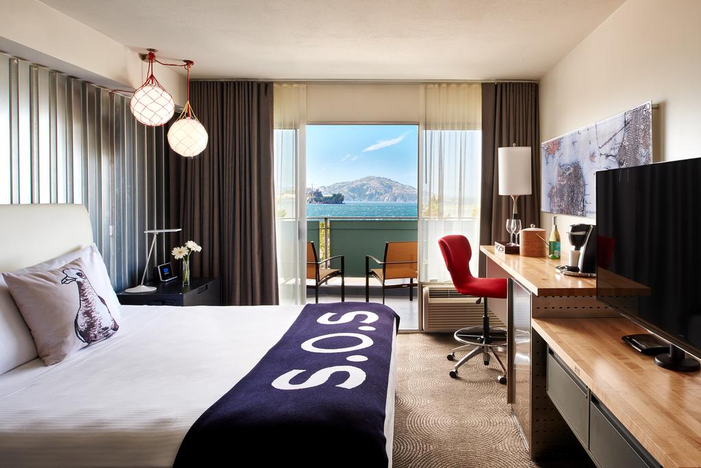 Photo of guest room at Hotel Zephyr in San Francisco. It is a very whimsical hotel which makes it where to stay with kids. Bed features a blue throw blanket with S.O.S in white lettering. An open balcony is seen with a view of blue water and a rocky shoreline in the distance. 