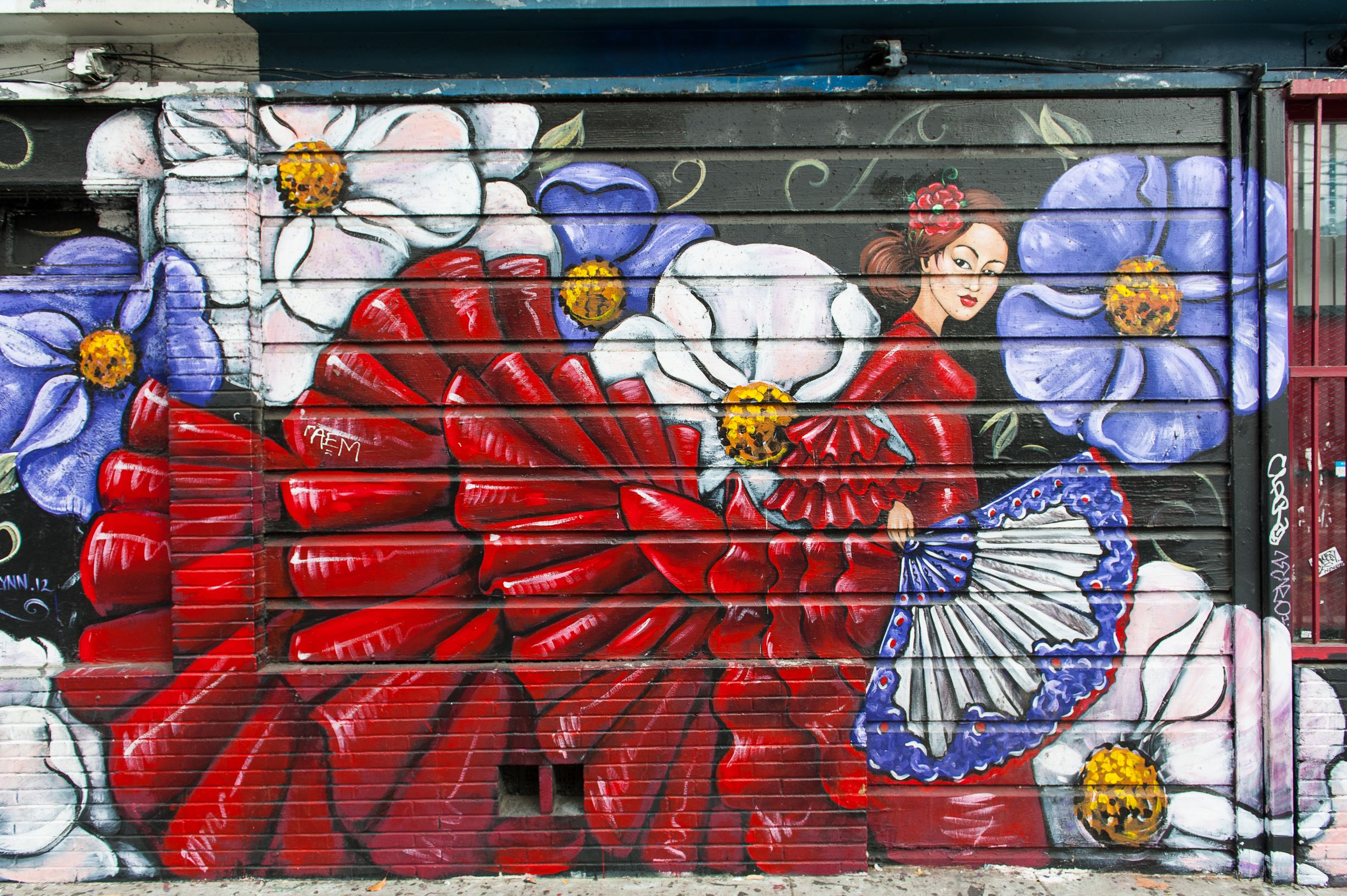 Photo of a colorful outdoor mural in the Mission District of San Francisco. Large white and purple flowers are painted around a woman in flamenco style red dress. 