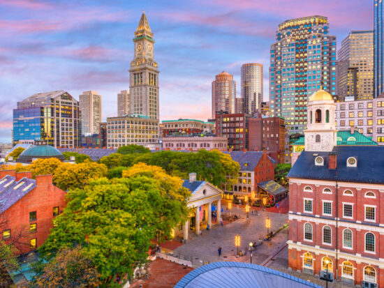 The skyline of Boston, Massachusetts at sunset. You can see very old buildings mixed with modern skyscrapers. The sky is blue, pink, and purple.