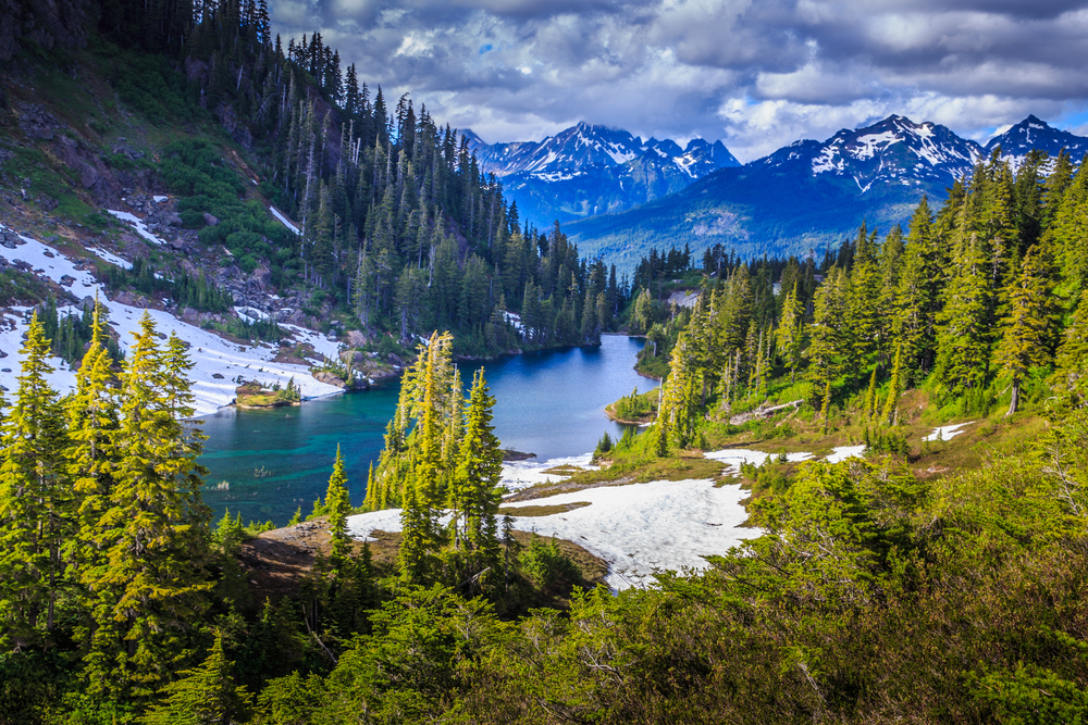 Blue lake in Montana with clouds, snow, and green trees in the foreground 
