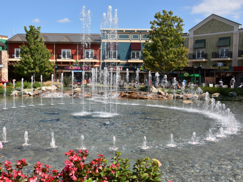 A view of the fountain show at The Island, Pigeon Forge. Spurts of water shoot up from sparkling water in a circle pattern. A line of charming shops cross the background. 