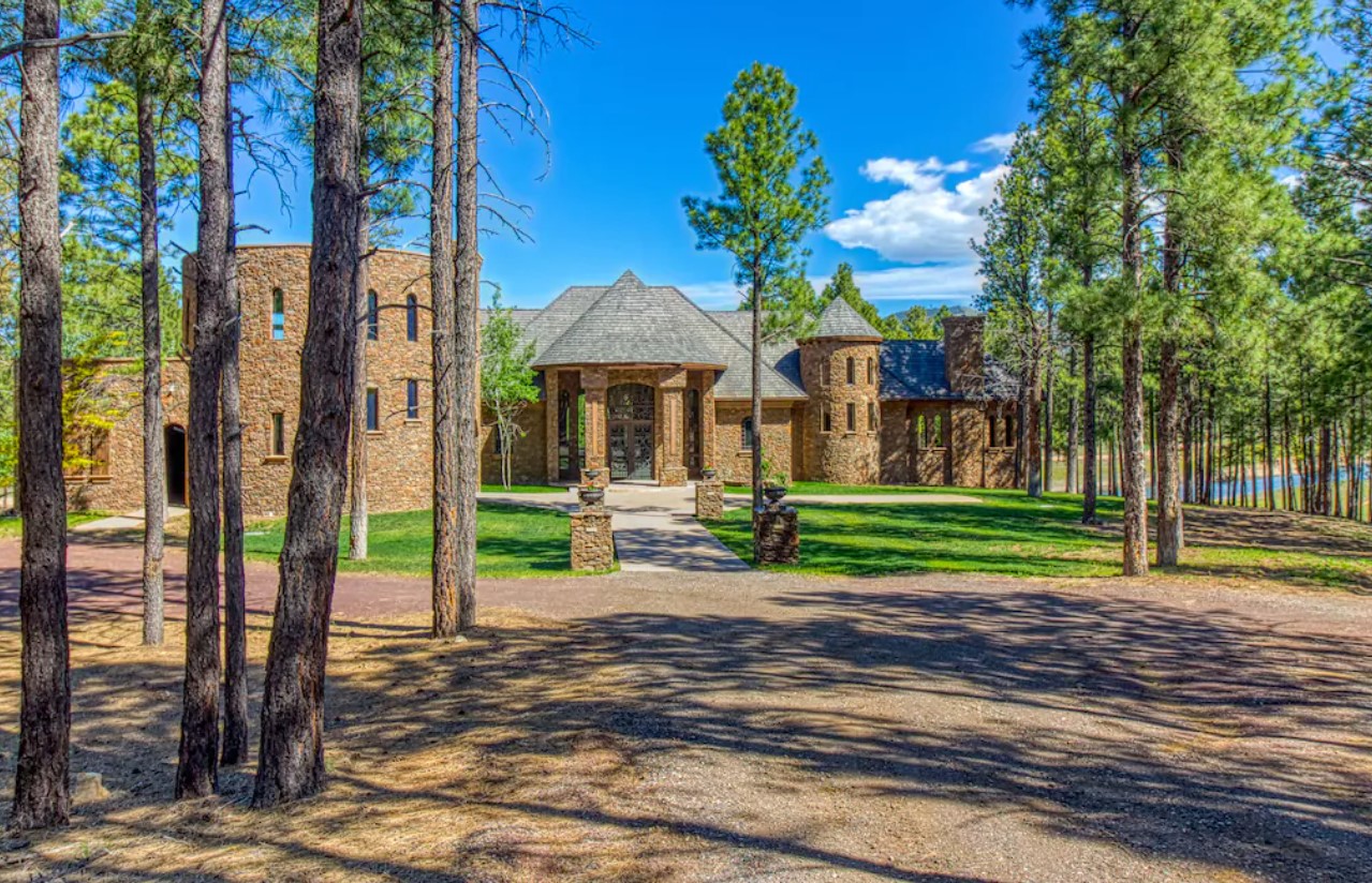 A large stone castle with turrets, a large and green front lawn, tall trees, on a sunny day