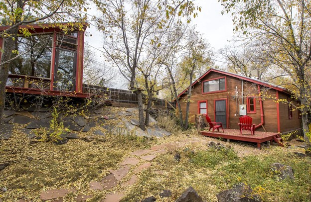 A cabin style home with red trim, a large deck with red chairs, and a glass walled studio across the backyard with grass, trees, and yellow flowers