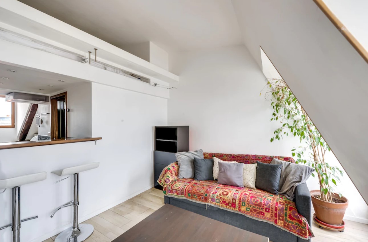 A charming studio with simple white walls, a small couch with a colorful quilt, large windows, and a tall palm tree in a pot.