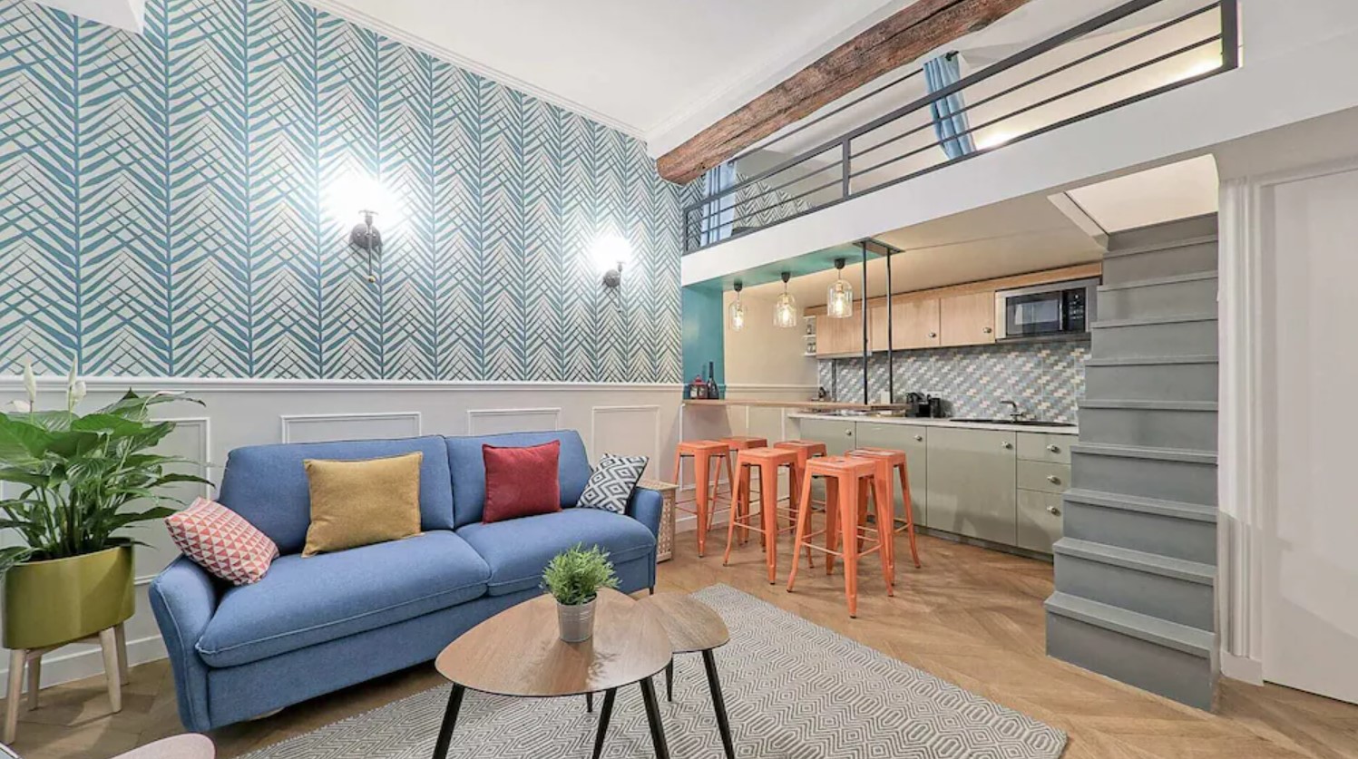 The inside of a colorful loft apartment in paris. It has blue and white wallpaper, a blue couch, peach bar stools, and a small kitchen.