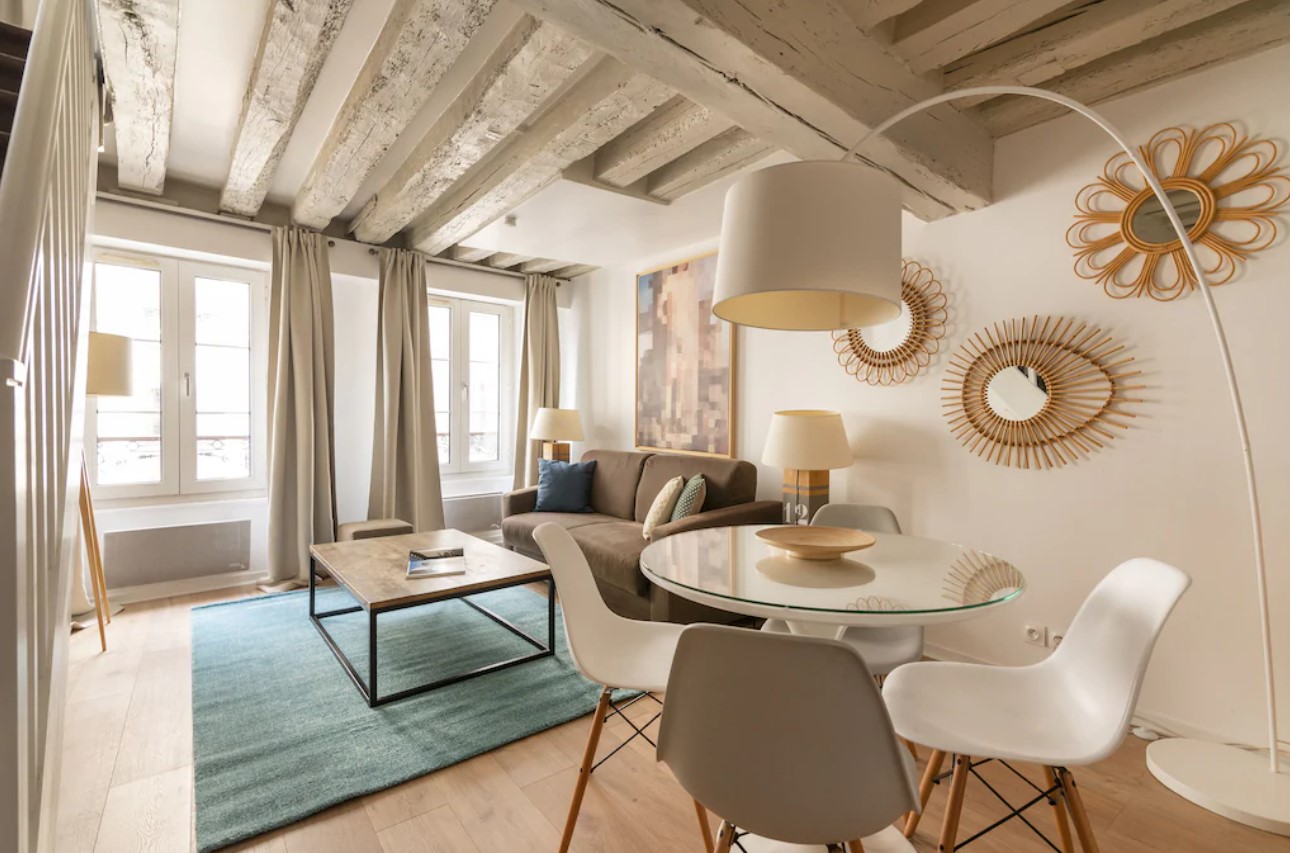 the living room of a simply decorated studio in paris. There are cream walls and painted wood beams, a small couch, a small table, and rattan mirrors on the wall. It's where to stay in paris. 