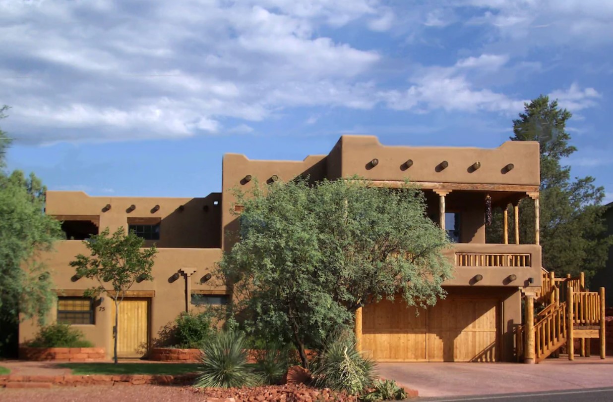 The exterior of a unique adobo style home made of brown clay, wood supports, and surrounded by trees on a sunny day