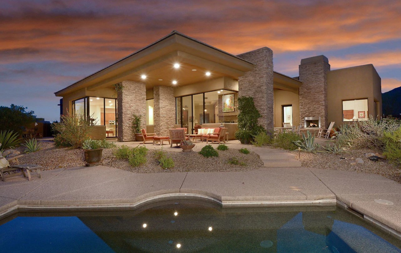 A large luxury modern home all lit up with a pool, large windows, a private patio, and desert plants at sunset