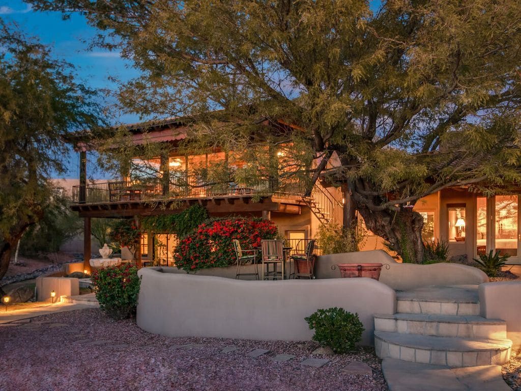 The exterior of a home lit up at twilight with a large loft and covered deck on the front of the house shaded by large trees with a large desert garden