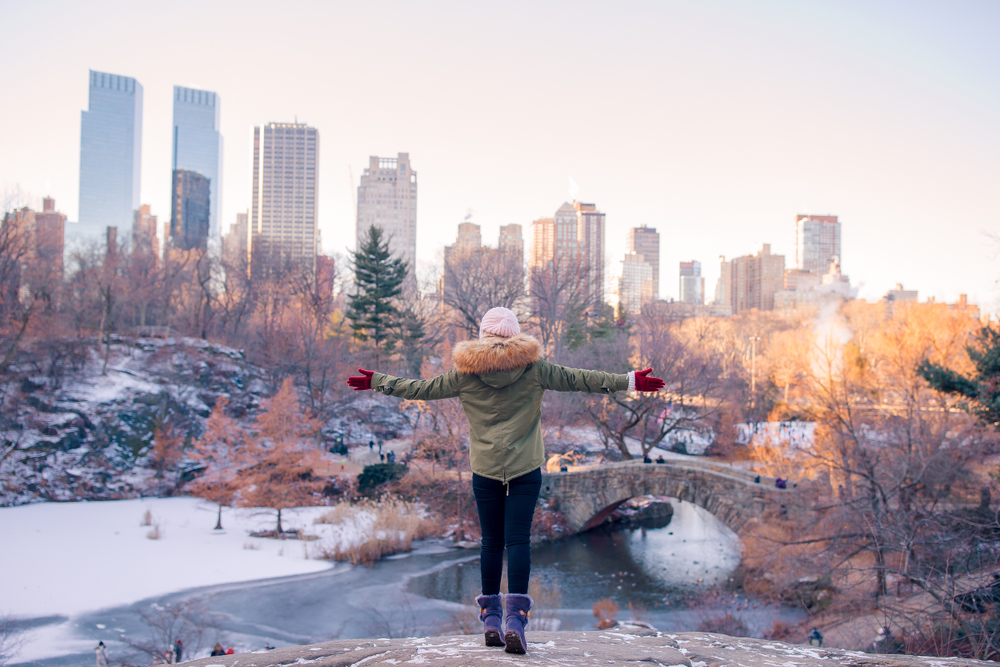 A woman with her arms held out bundled in layers in front of New York skyline in a snowy Central Park.