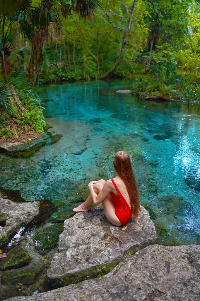 Photo of young woman in a red swimsuit sitting on a rock looking at the spring water in Florida.