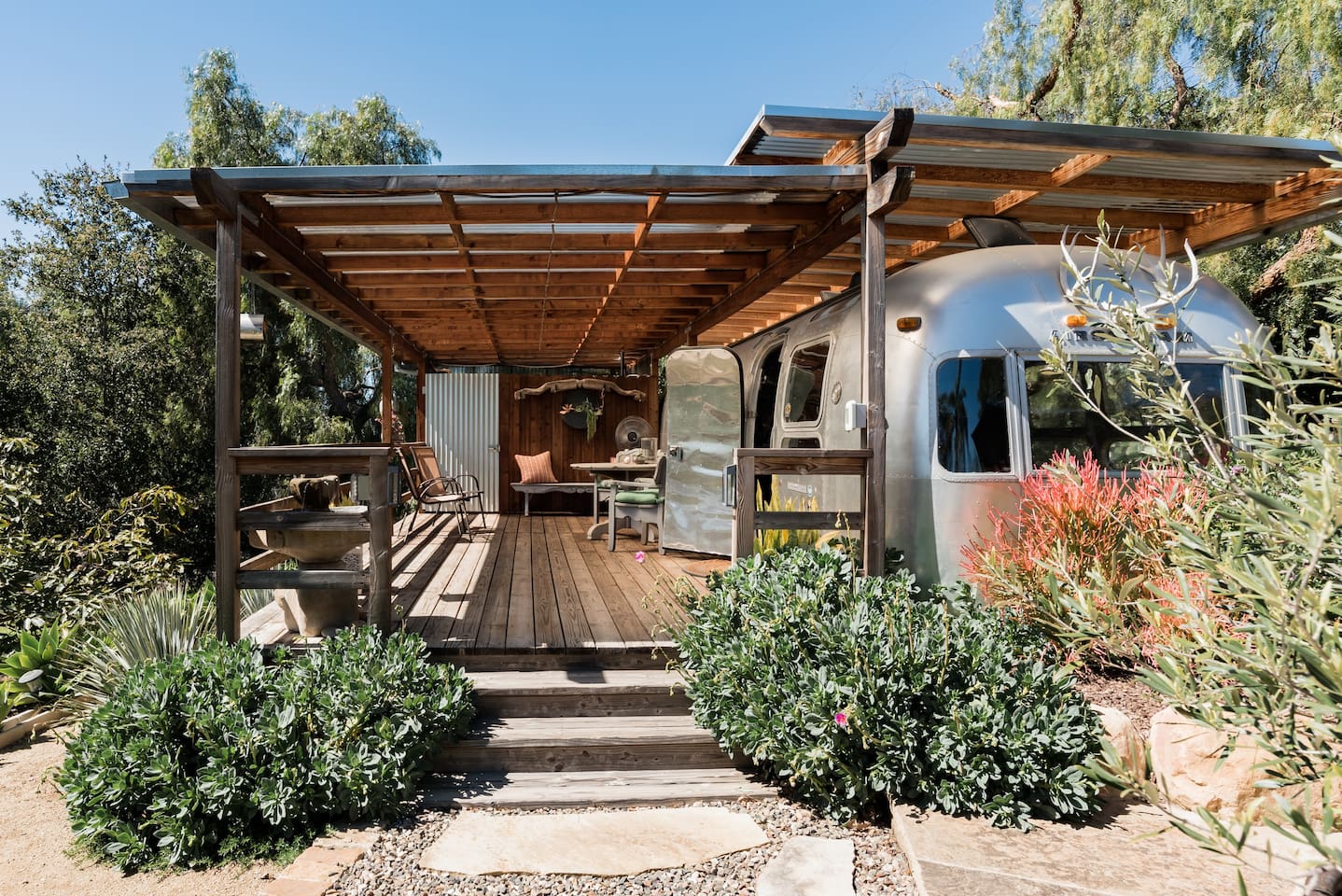 Cool Airbnb in California... 1974 Airstream, with a wooden deck and awning attached, surrounded by plants and trees.