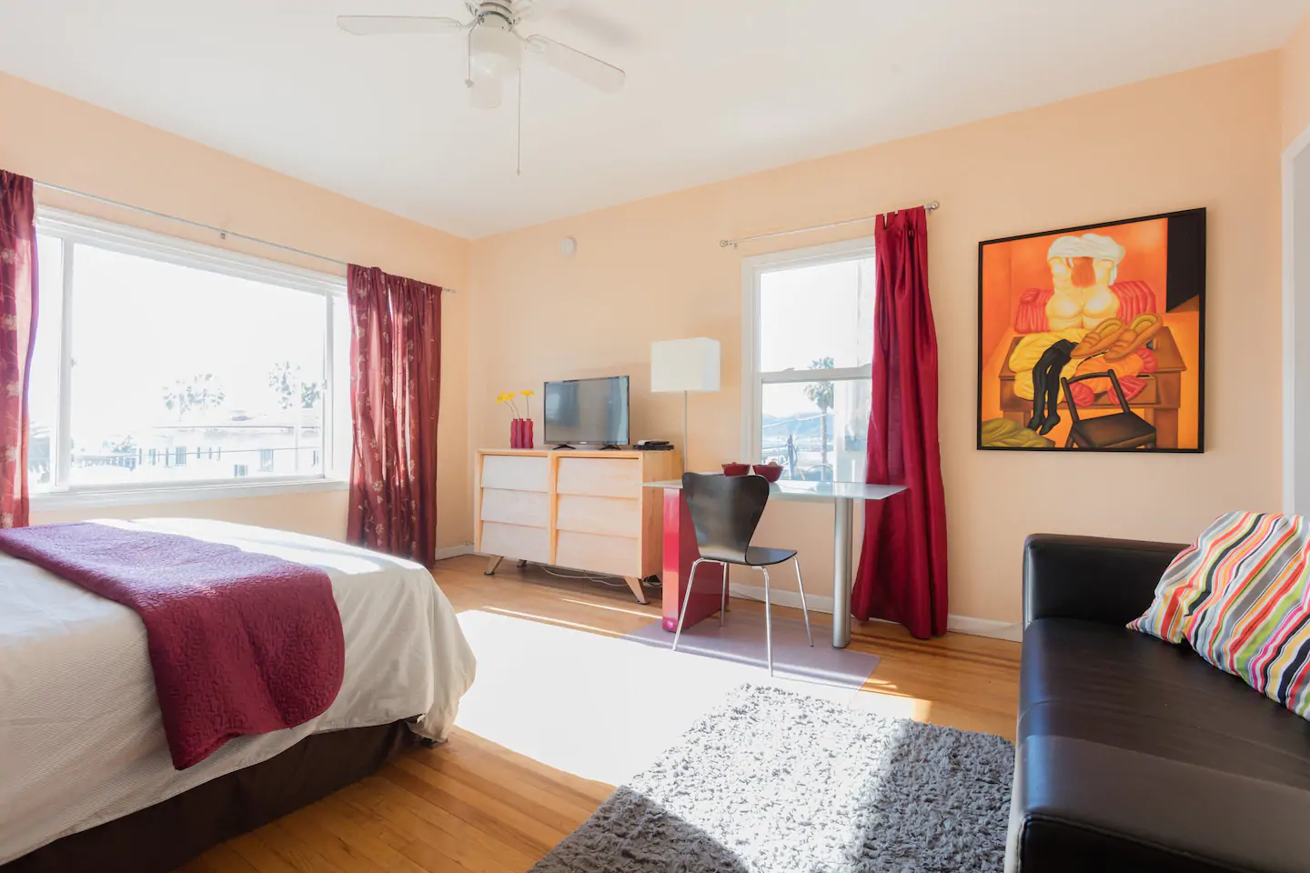 A well-lit bedroom in a studio Airbnb in California, with a bed, dresser, desk, couch, and bright red curtains.