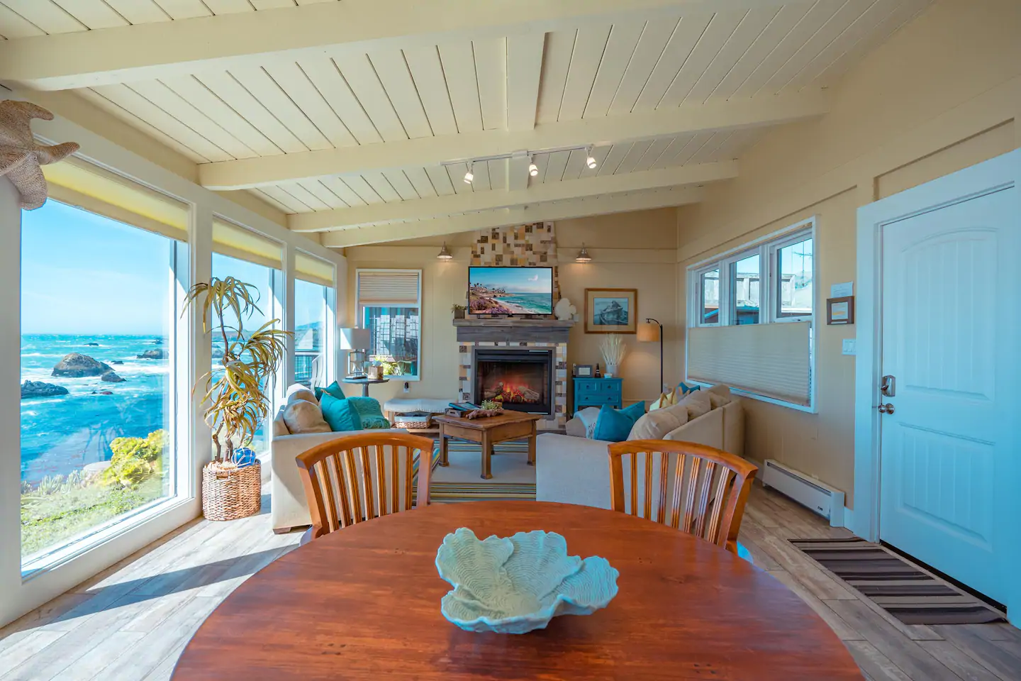 Airy, light, cozy beach house living area, with table, chairs, sofa seats, a fire place, and ocean view. Airbnb in California.