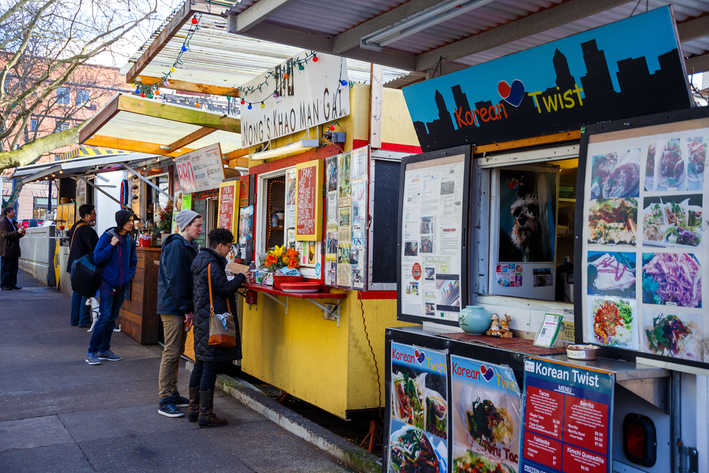 a line of food trucks in Portland, Oregon. The visible signs say "Korean Twist" "Nong's Khao Man Gai" and "BBQ" eating at food trucks is one of the best things to do in Portland, Oregon