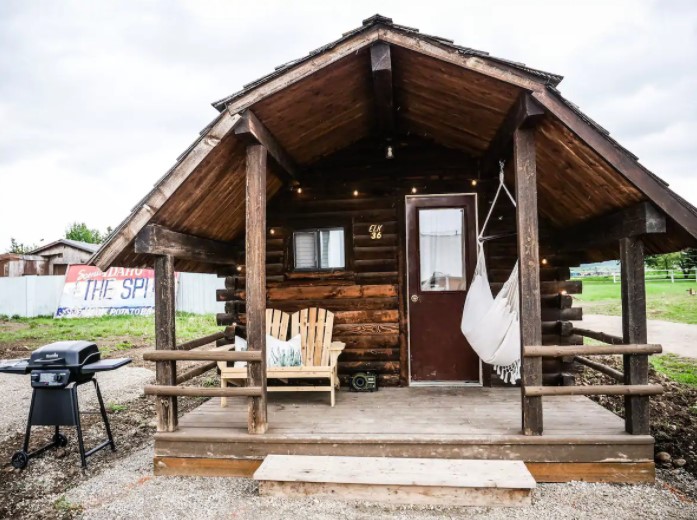 Front view of a small dark wood cabin with a hammock and Adirondack seats on a small porch