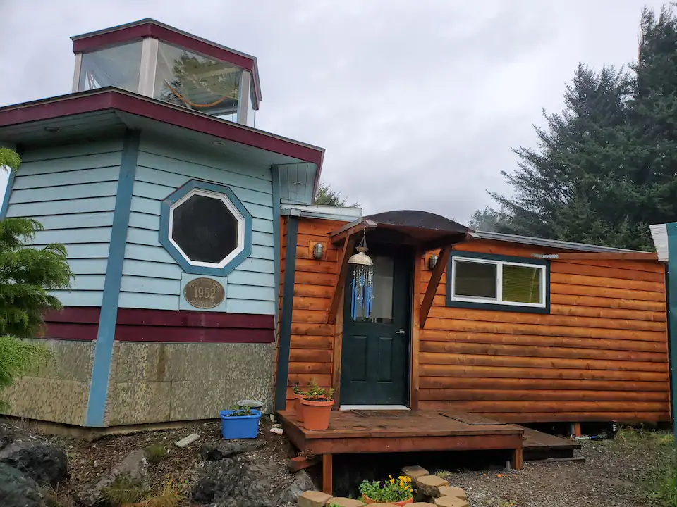 Exterior picture of the lighthouse cottage, one of the best cabins in Oregon. The cabin is half traditional wooded cabin and has an end cap that is a rounded two story blue and red lighthouse. 