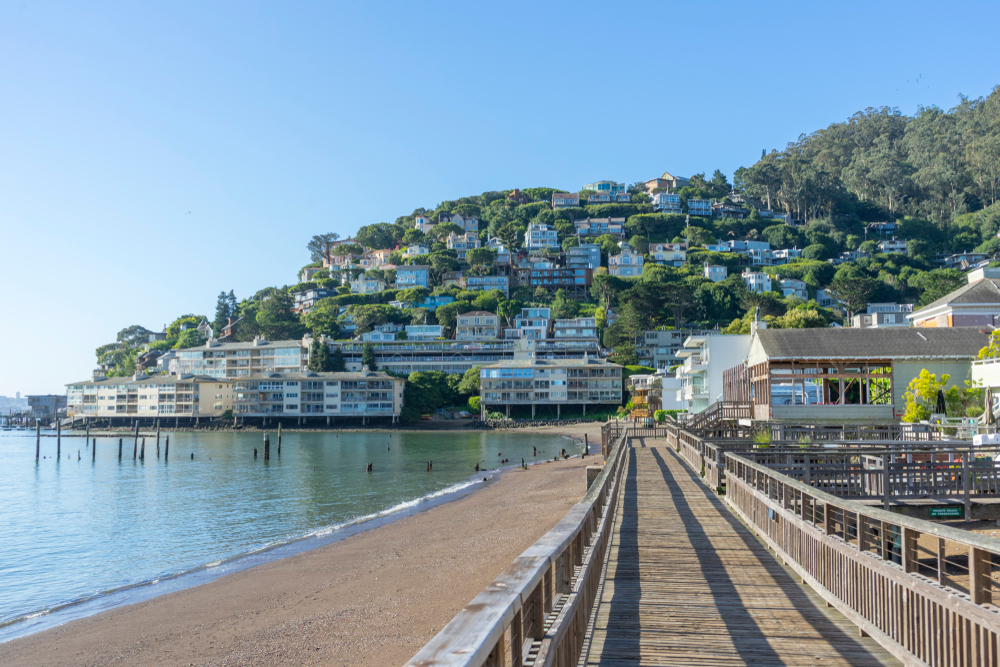 Looking down a narrow boardwalk next to the beach with the houses on a hill in the distance in Sausalito California