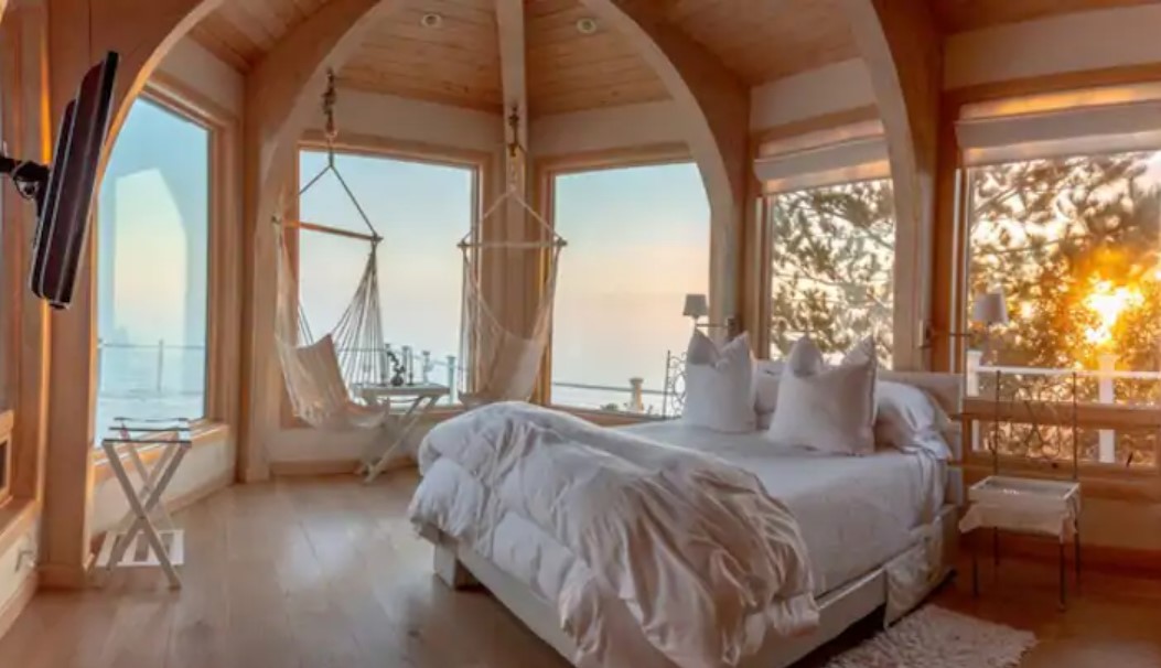 A queen-sized bed in a room full of windows that looks out to the ocean with two hammock seats inside