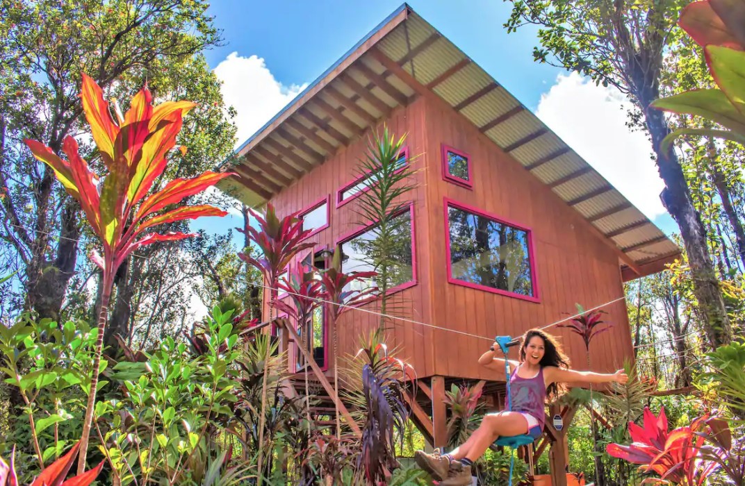 A woman using a small zip line outside of a tiny home treehouse surrounded by tropical plants one of the best airbnbs in Hawaii