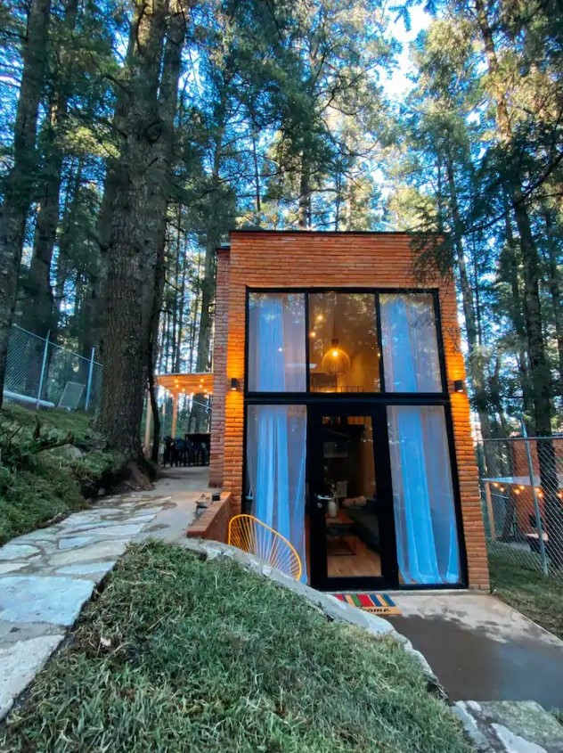 The outside of a brick cabin in the forest in Mexico with a small patio and floor to ceiling windows Airbnbs in Mexico