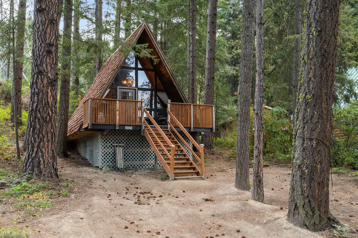 A frame cabin with a wall of windows surrounded by evergreen trees. There are about 10 steps leading to the spacious front deck.  