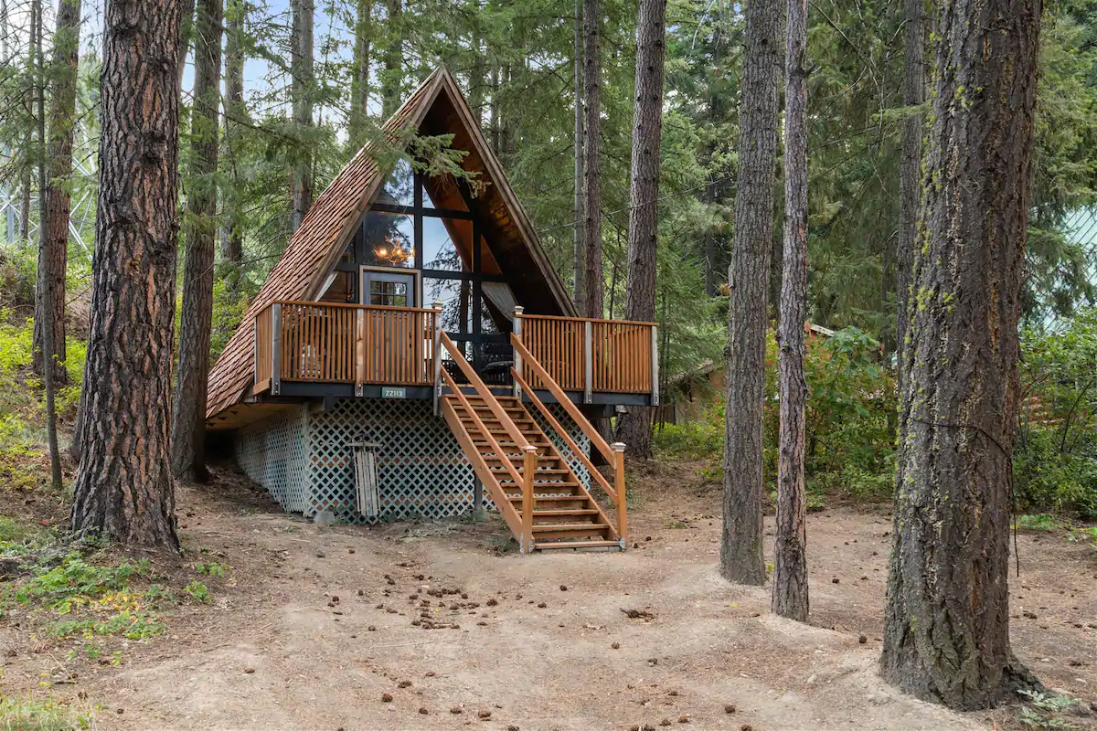 A frame cabin with a wall of windows surrounded by evergreen trees. There are about 10 steps leading to the spacious front deck.