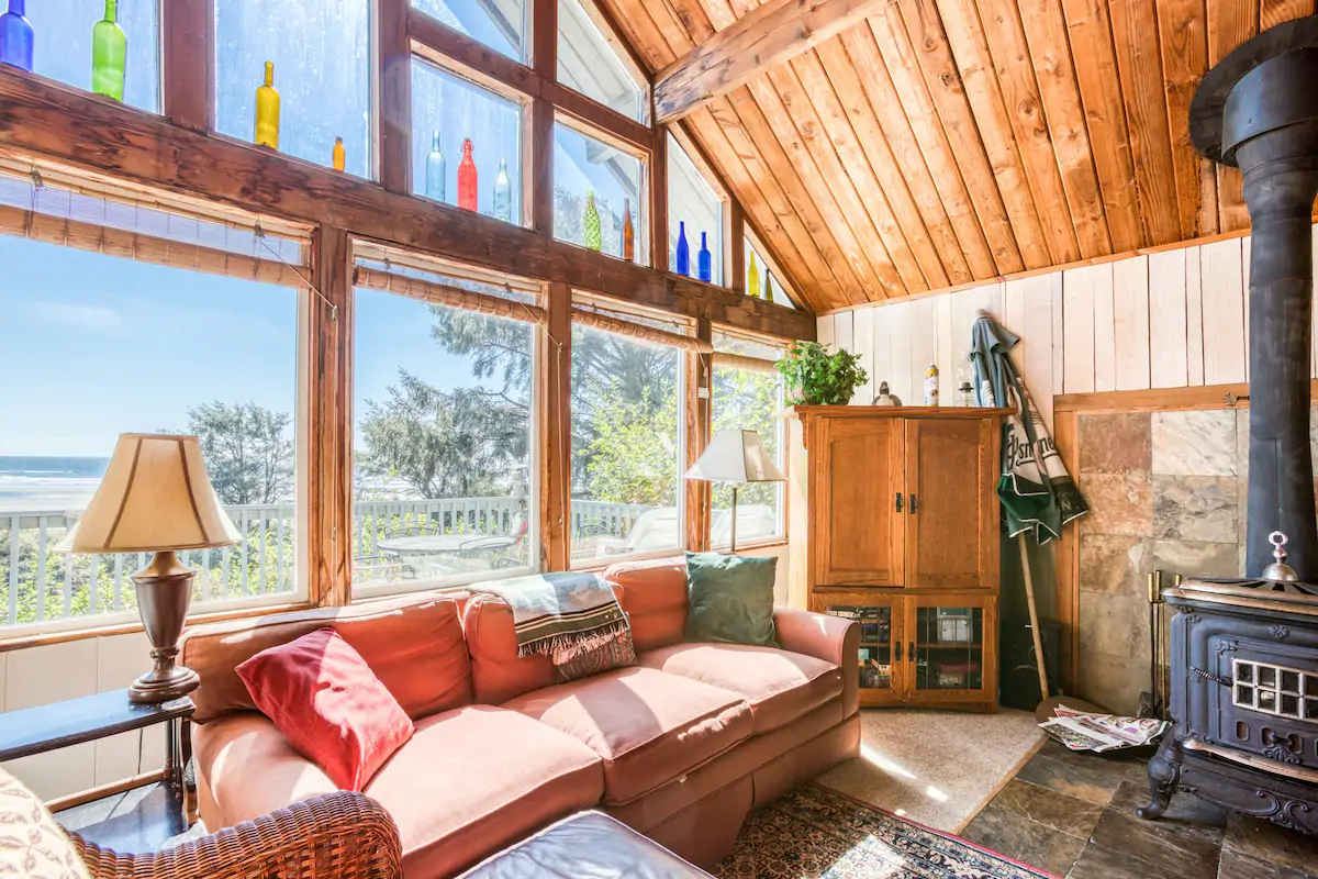 Cozy terracotta colored living room couch by a woodstove. There is a wall of windows behind the couch looking out at a large deck, and beyond that a beautiful beach. 