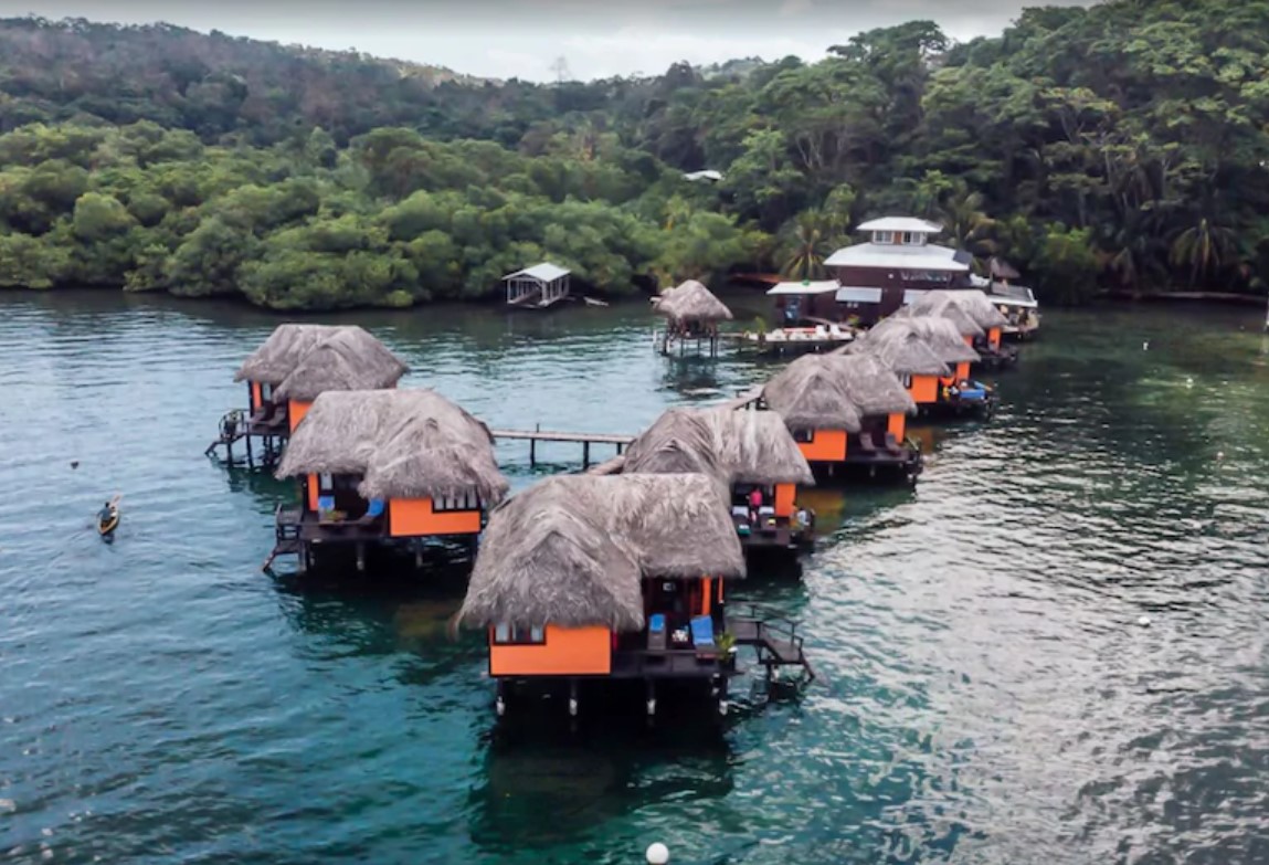 An aerial view of seven bright orange overwater bungalows with natural thatch rooves in the Caribbean