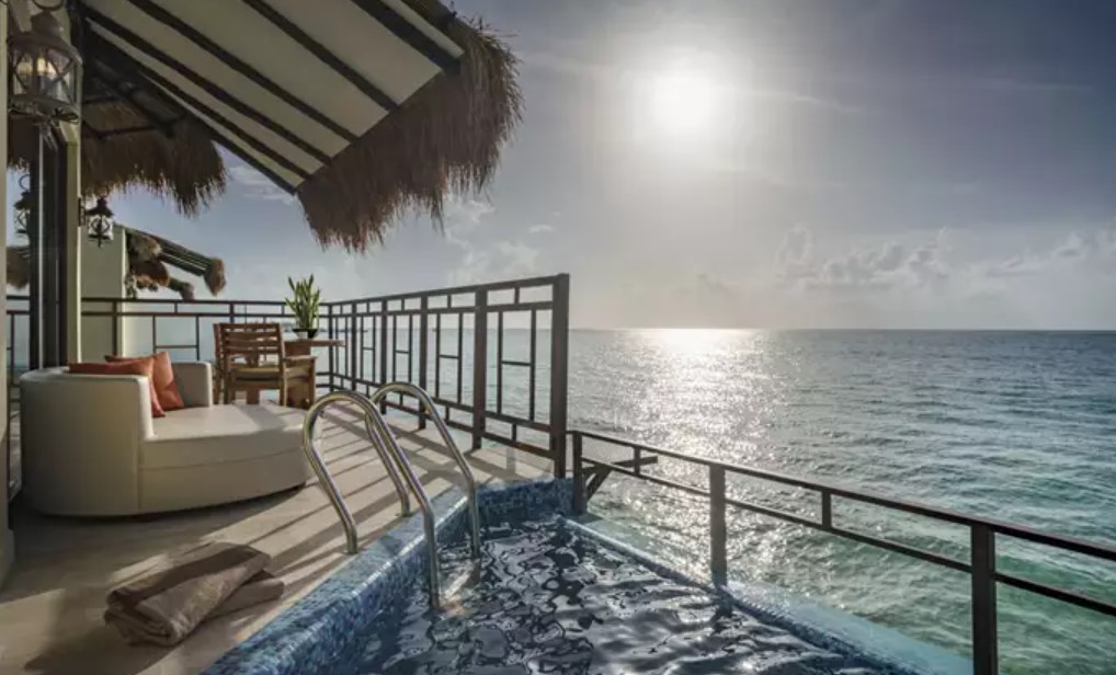 The deck of an overwater bungalow with a sun lounger couch, a bistro set, and a hot tub.