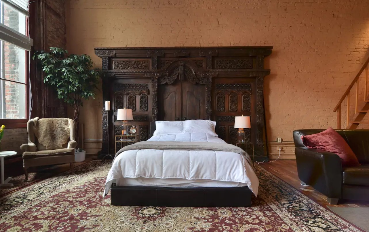 A large studio space with painted brick walls, large windows, and a large ornate wooden headboard behind a queen-sized bed in Seattle 