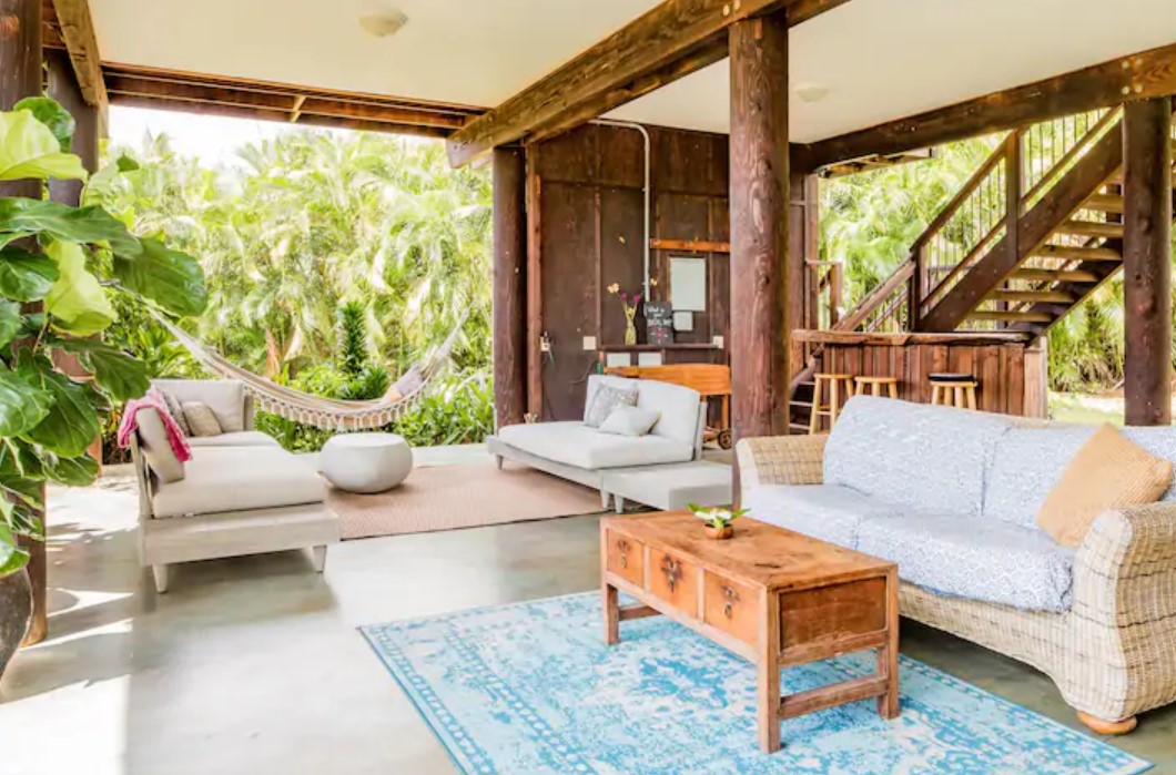 Looking at an open air living space with two couches, a hammock, and a bar space surrounded by tropical greenery