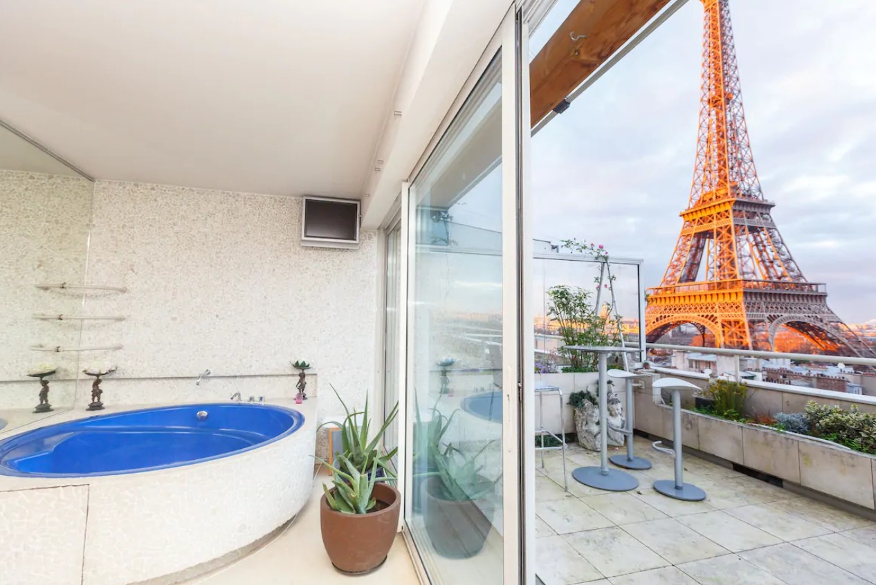 A hot tub behind the glass of a balcony and the balcony is large with seating and amazing views of the Eiffel Tower