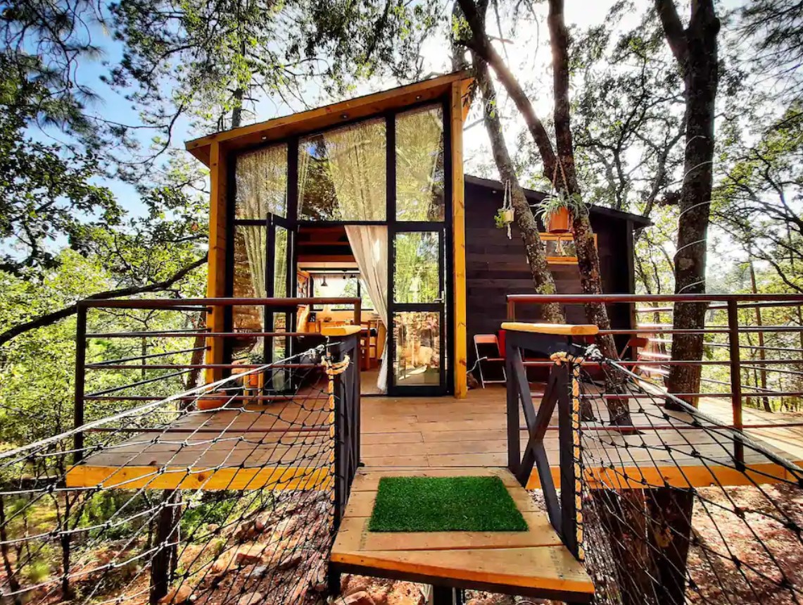 The exterior view of an elevated treehouse in the woods in Mexico with a large deck and floor to ceiling windows