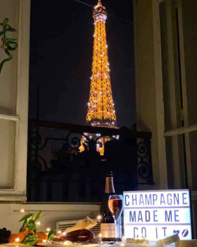 The view of the Eiffel Tower at night through and open window with a small table set up in front of it with champagne and food on it