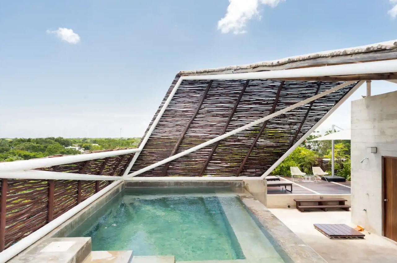 A large roof top pool with a bamboo awning over it and a wooden sunbathing deck