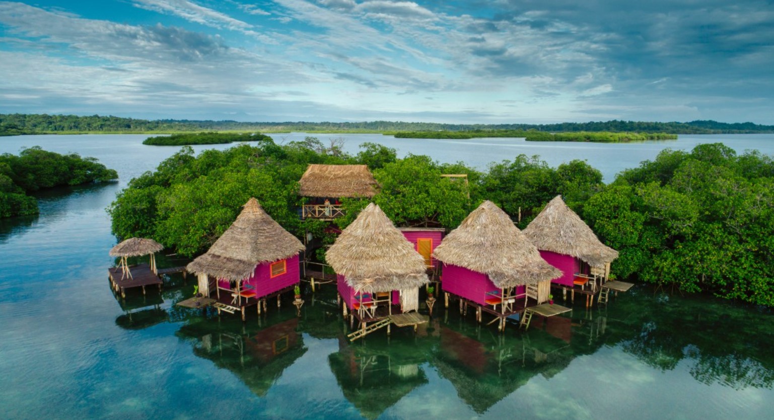 An aerial view of bright pink overwater bungalows in the caribbean with thatch rooves surrounded by jungle and the ocean