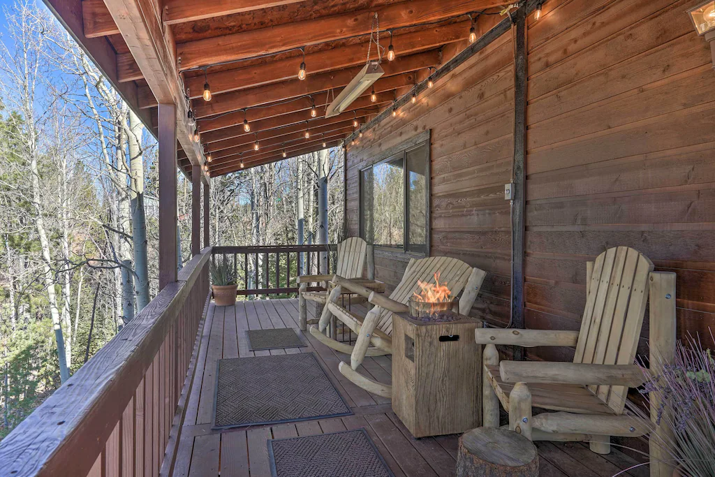 Cabin shwing large deck with view. There are wooden chairs on the deck and a fire. 